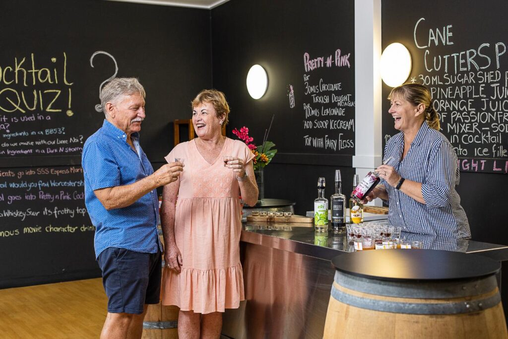 Two older adults sample drinks at a bar while a bartender pours liquor. Chalkboards with cocktail recipes and a quiz are visible in the background.
