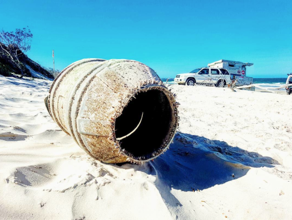 A large rusty metal drum lies on a sandy beach with a white vehicle and the ocean in the background under a clear blue sky.