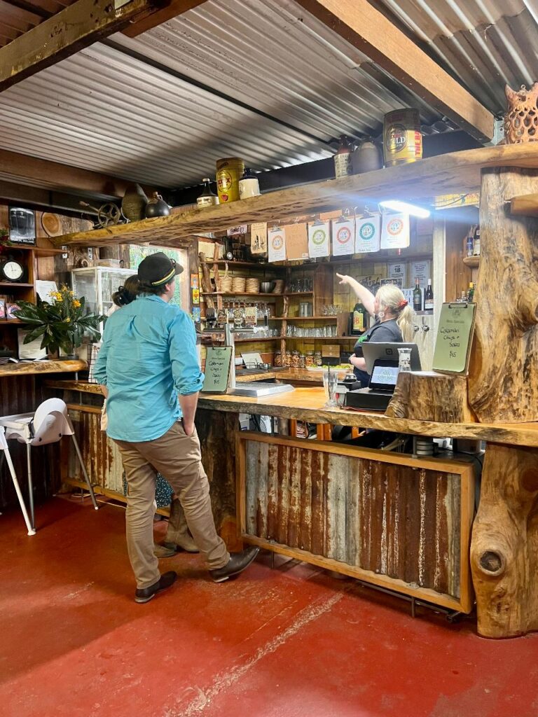A person stands at a rustic wooden bar counter while an employee behind the counter points at items on a shelf; shelves display jars, bottles, and decor.