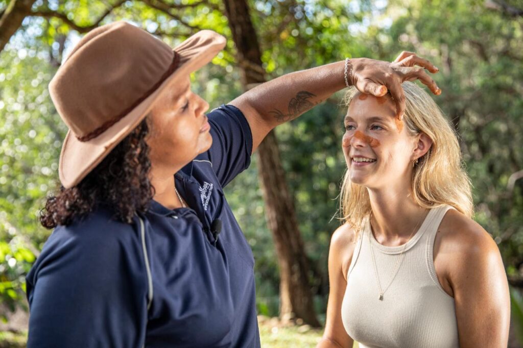 A woman in a hat applies orange paint to another woman's face outdoors in a forested area.