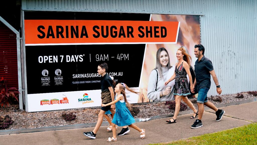 A family of four walks past a building with a large sign for Sarina Sugar Shed, which states it is open 7 days a week from 9am to 4pm.