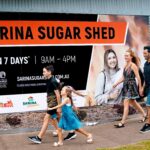 A family of four walks past a building with a large sign for Sarina Sugar Shed, which states it is open 7 days a week from 9am to 4pm.