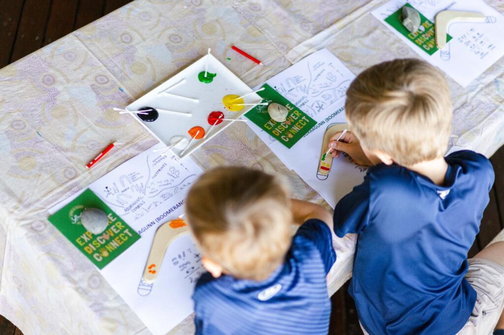 Two children sit at a table, painting colorful drawings on paper with paint and brushes; paint pots and art supplies are scattered on the table.