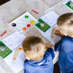 Two children sit at a table, painting colorful drawings on paper with paint and brushes; paint pots and art supplies are scattered on the table.