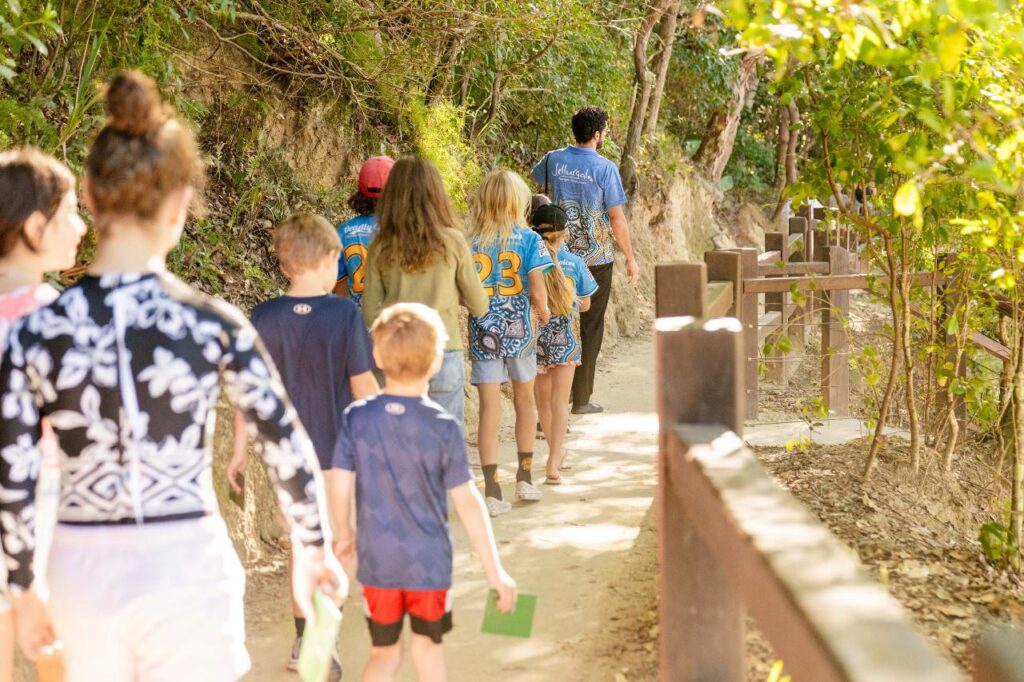A group of children and adults walk along a dirt path through a wooded area, some wearing matching blue shirts with numbers on the back.