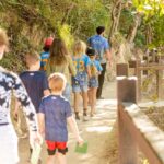 A group of children and adults walk along a dirt path through a wooded area, some wearing matching blue shirts with numbers on the back.