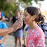 A person applies white paint to a smiling girl's face outdoors, with other people and trees visible in the background.