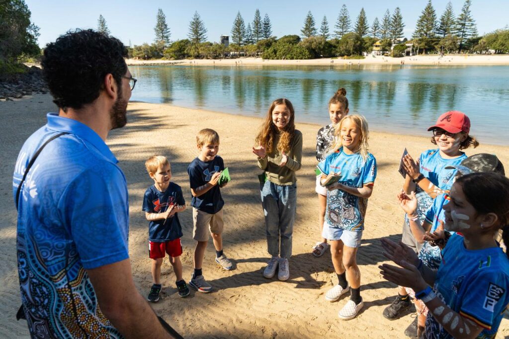 A group of children and an adult stand in a circle on a sandy riverbank, smiling and clapping during an outdoor activity on a sunny day.