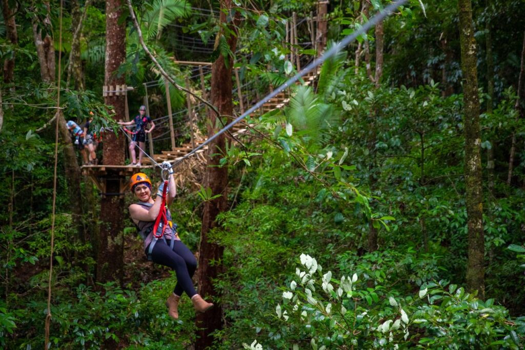A person wearing a helmet and harness rides a zipline through a lush green forest, with others waiting on a wooden platform in the background.