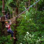 A person wearing a helmet and harness rides a zipline through a lush green forest, with others waiting on a wooden platform in the background.