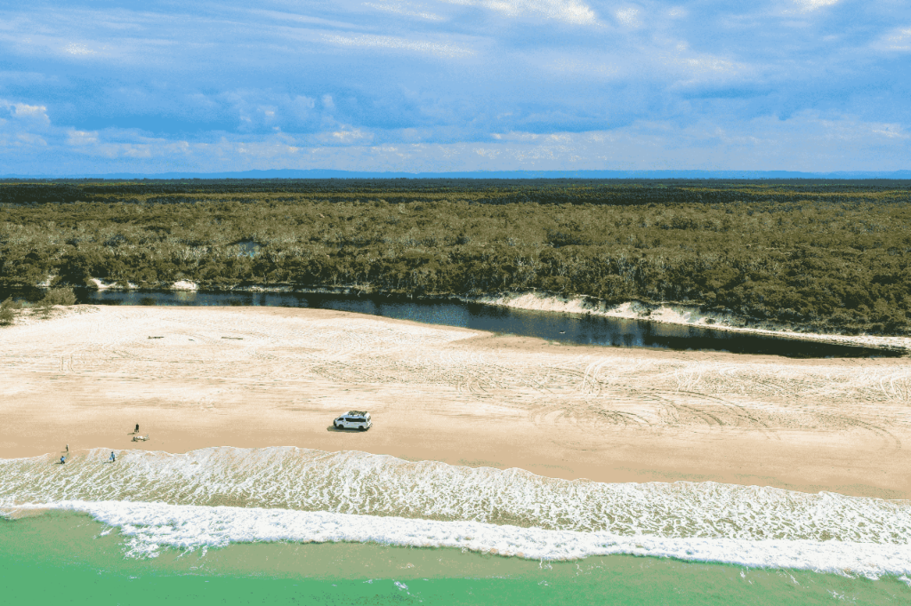 Aerial view of a sandy beach with a parked vehicle near the shore, a few people nearby, a narrow river, and a forested area in the background under a partly cloudy sky.