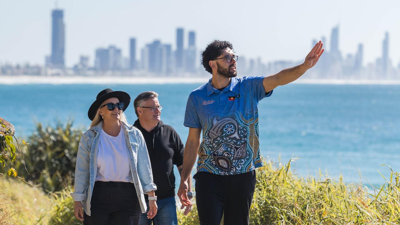 Three people walk along a coastal path with city buildings in the background; one man gestures while talking to the others.