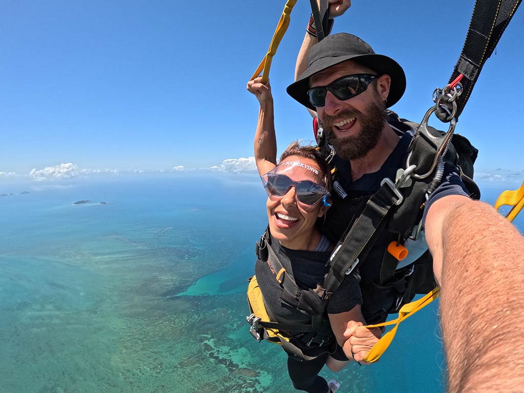 Two people tandem skydiving high above a clear blue ocean, smiling at the camera, with islands visible in the distance.