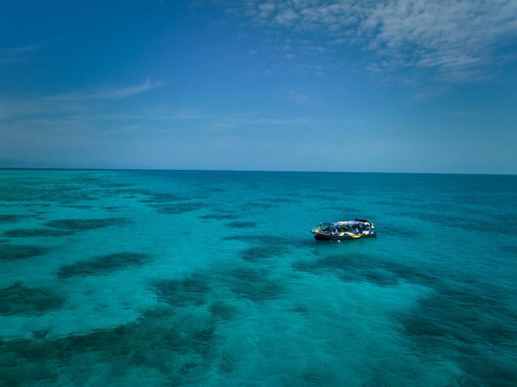 A small boat floats on clear turquoise water with visible coral reefs under a blue sky with scattered clouds.