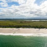 Aerial view of a sandy beach bordered by green ocean waves, with a forested area and distant mountains under a partly cloudy sky.