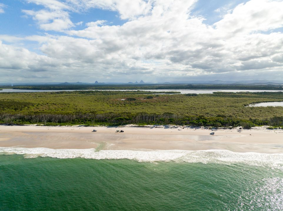 Aerial view of a sandy beach bordered by green ocean waves, with a forested area and distant mountains under a partly cloudy sky.