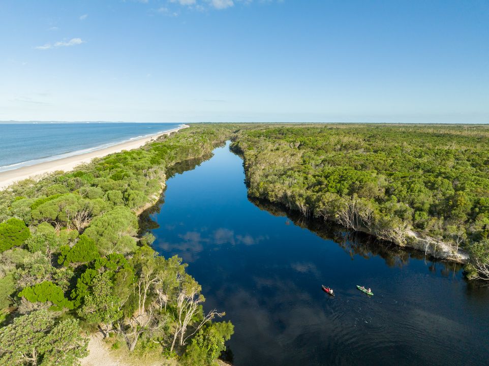 Aerial view of a river flowing through dense green forest toward a sandy beach, with two kayaks paddling on the water under a clear blue sky.
