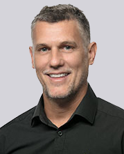 Smiling man with short gray hair and a trimmed beard, wearing a black collared shirt, posed against a plain light gray background.