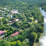 Aerial view of a small town with buildings surrounded by dense greenery, adjacent to a river with a bridge crossing over it.