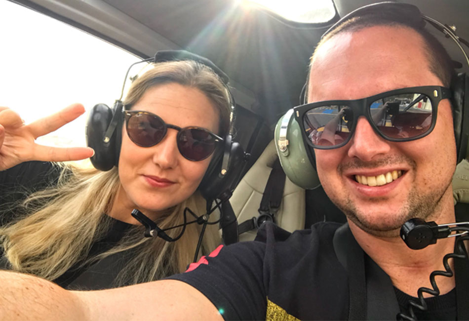 Two people wearing sunglasses and headsets sit inside a helicopter; one is making a peace sign, both are smiling.