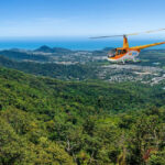 A yellow helicopter flies over a green forested hillside with a coastal city and ocean visible in the background under a clear blue sky.