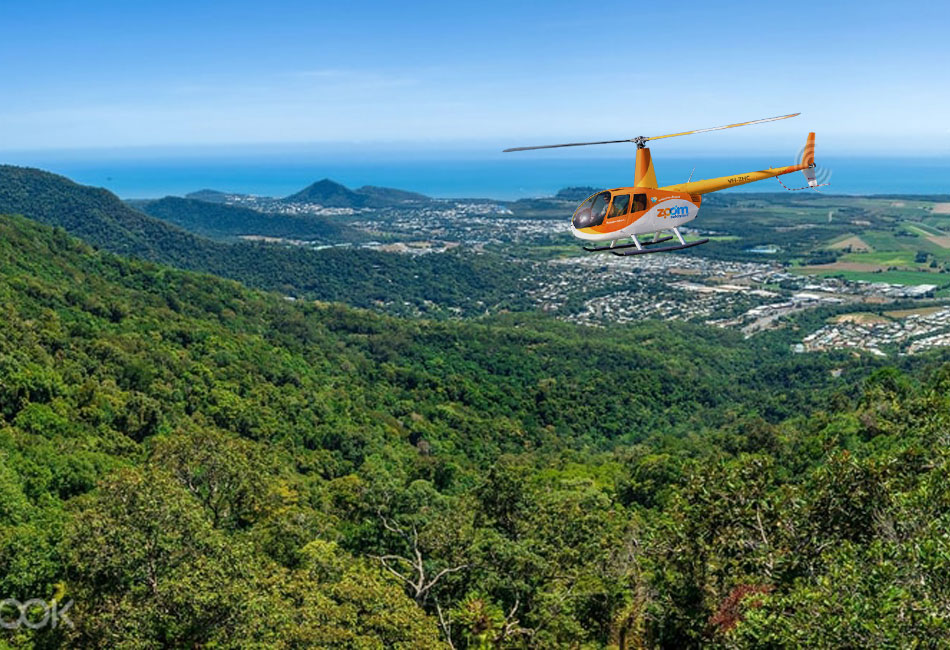 A yellow helicopter flies over a green forested hillside with a coastal city and ocean visible in the background under a clear blue sky.