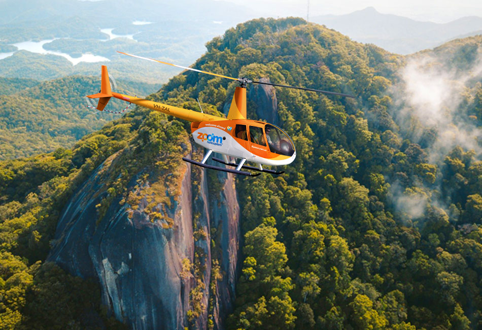 An orange and white helicopter with "Zoom" branding flies over a lush green mountainous landscape with rocky cliffs and trees.