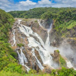 A large, multi-tiered waterfall flows over rocky cliffs surrounded by dense green forest under a partly cloudy sky.