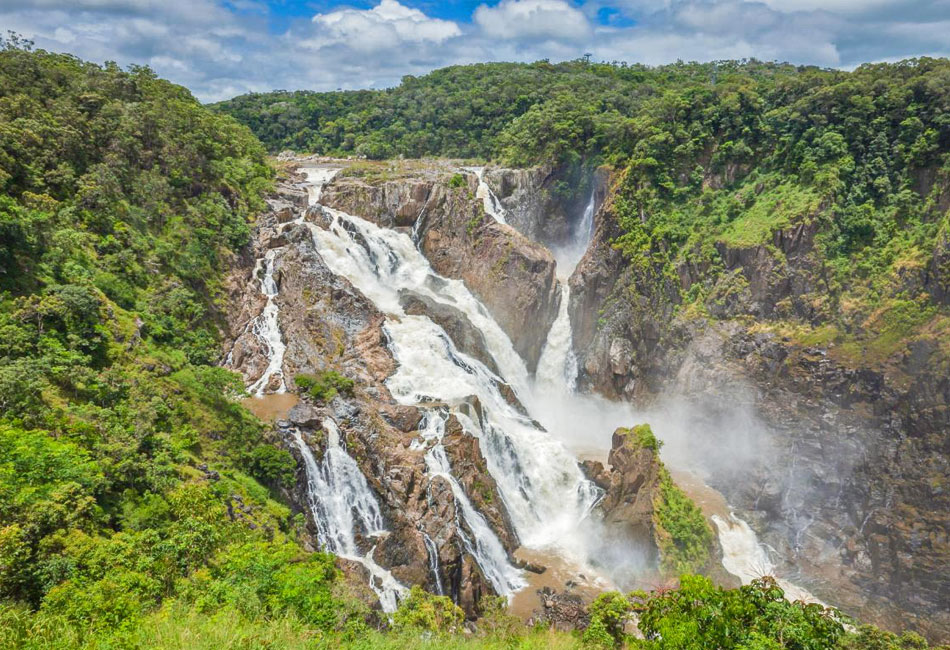 A large, multi-tiered waterfall flows over rocky cliffs surrounded by dense green forest under a partly cloudy sky.