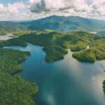 Aerial view of a forested landscape with a winding lake, surrounded by green hills and mountains under a partly cloudy sky.