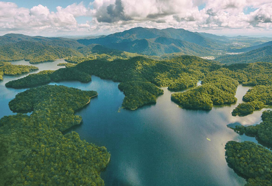 Aerial view of a forested landscape with a winding lake, surrounded by green hills and mountains under a partly cloudy sky.