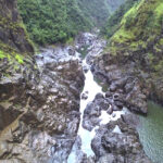 Rocky river gorge with steep, green-clad cliffs and patches of water flowing between large rugged rocks.