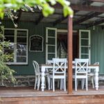 A wooden deck with a white dining table and chairs sits outside a green corrugated metal house with open French doors and a chalkboard sign on the wall.