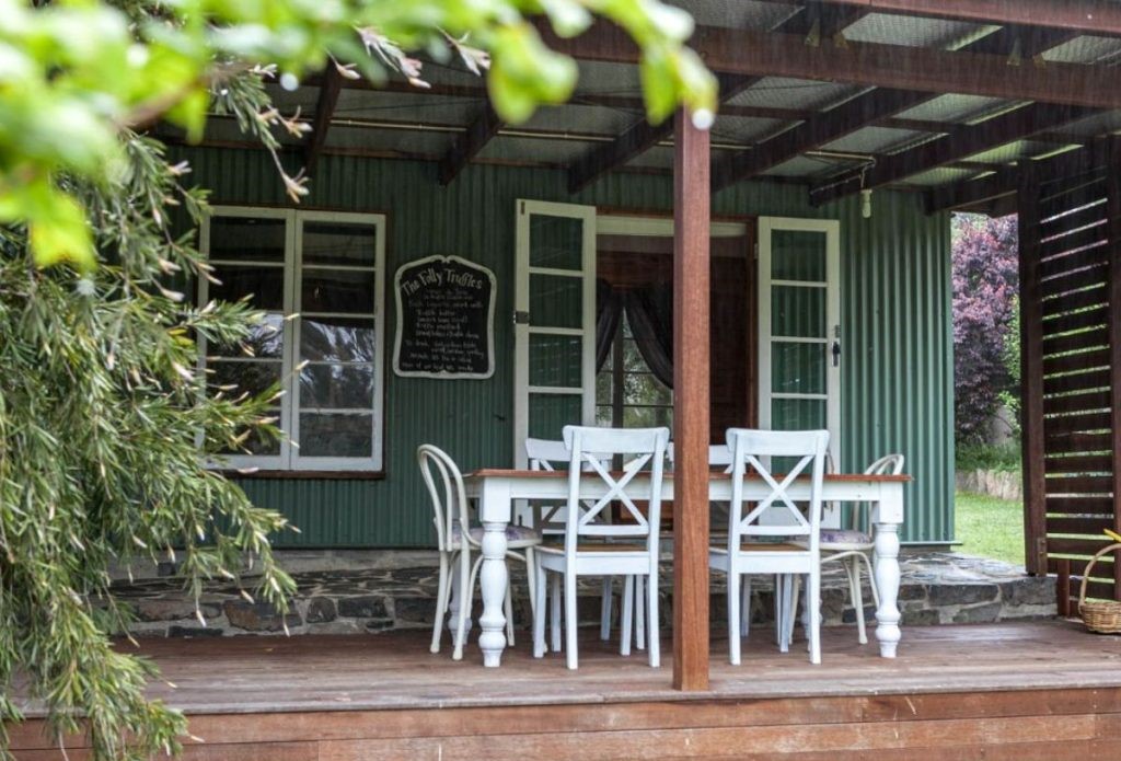 A wooden deck with a white dining table and chairs sits outside a green corrugated metal house with open French doors and a chalkboard sign on the wall.