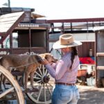 A woman in a hat stands beside a chained dog on a wooden wagon, gently holding the dog's face in an outdoor rural setting.