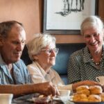Three older adults sit at a table with tea and scones, one woman pours tea while the others smile and enjoy the conversation.