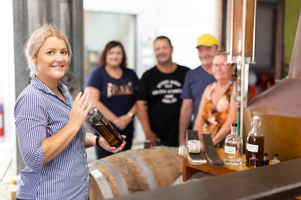 A woman holds a bottle and smiles at the camera in a distillery, with four people standing and smiling in the background near barrels and equipment.