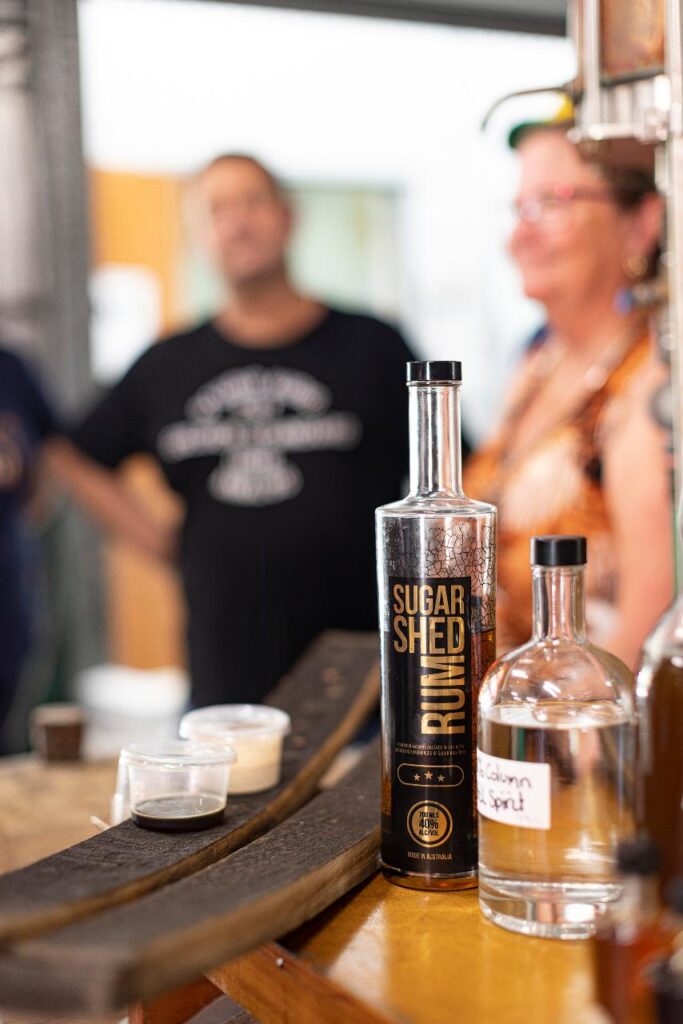 Bottles of rum and spirits on a counter with two tasting cups; two people stand blurred in the background.