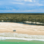 A car is parked on a sandy beach near the shoreline, with a river and forest in the background under a partly cloudy sky.