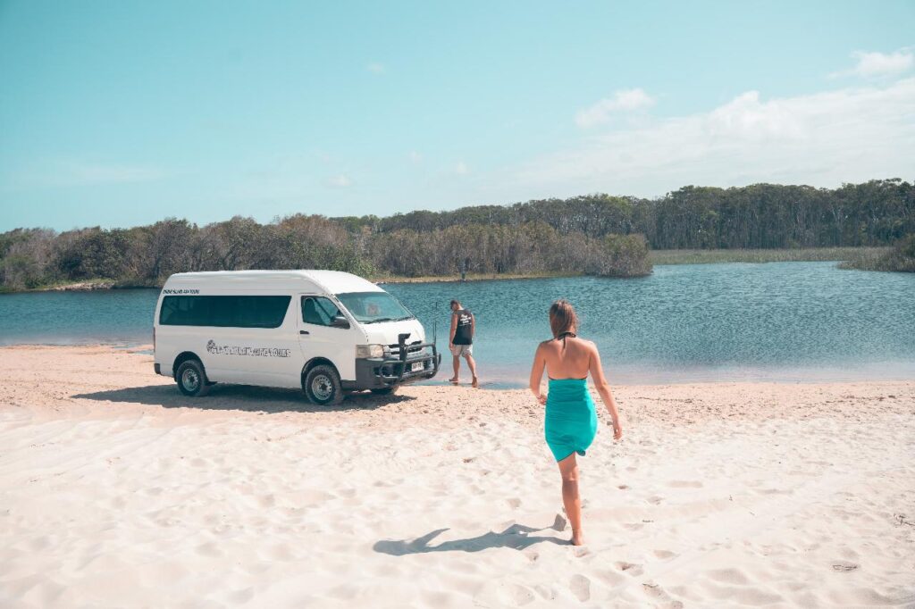 A woman in a turquoise dress walks on sand toward a person standing by a white van near a lake bordered by trees.