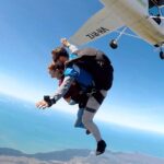 Two people tandem skydiving with an instructor, free-falling from an airplane over a coastal landscape under a clear blue sky.