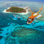 An orange helicopter flies over turquoise waters and coral reefs near a small, green island with a dock and several boats.