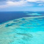 Aerial view of clear turquoise water and coral formations in a large reef system under a blue sky with sparse clouds.