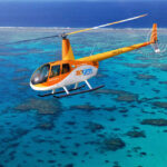 An orange and white Zoom Helicopters chopper flies over clear blue water and coral reefs.