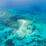 Aerial view of a small sandy island surrounded by turquoise and deep blue ocean waters with scattered coral reefs.