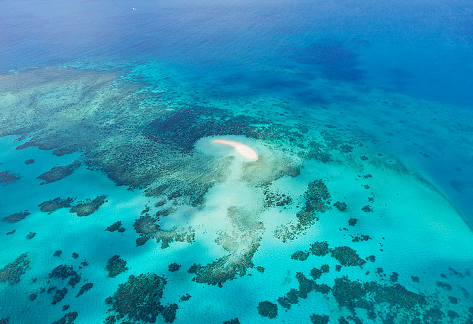 Aerial view of a small sandy island surrounded by turquoise and deep blue ocean waters with scattered coral reefs.