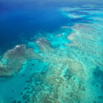 Aerial view of a coral reef in clear turquoise ocean water, showing shallow and deep areas with visible patches of coral formations.
