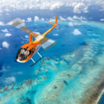 An orange helicopter flies over a turquoise ocean with coral reefs and shallow waters visible below, under a partly cloudy sky.