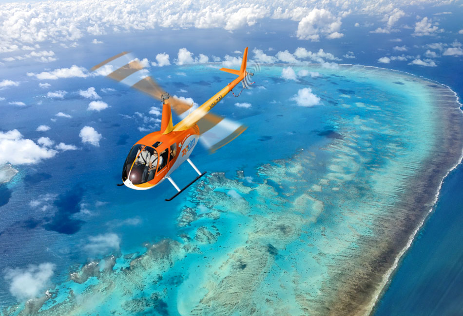 An orange helicopter flies over a turquoise ocean with coral reefs and shallow waters visible below, under a partly cloudy sky.