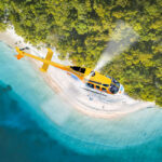 A yellow helicopter flies above a secluded beach surrounded by turquoise water and dense green forest.